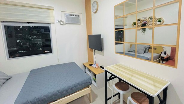 A bedroom with a bed, a desk, two chairs, a mirror, a clock, and a window with a view of a cityscape. - City Comfort at Urban Deca Towers Cubao (Quezon City)