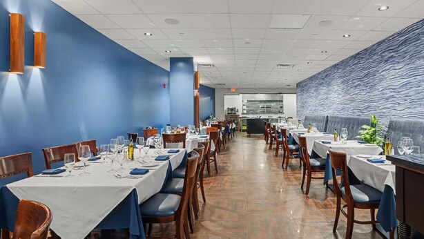 A restaurant with multiple tables and chairs, a kitchen in the background, and a blue wall with a wavy pattern.