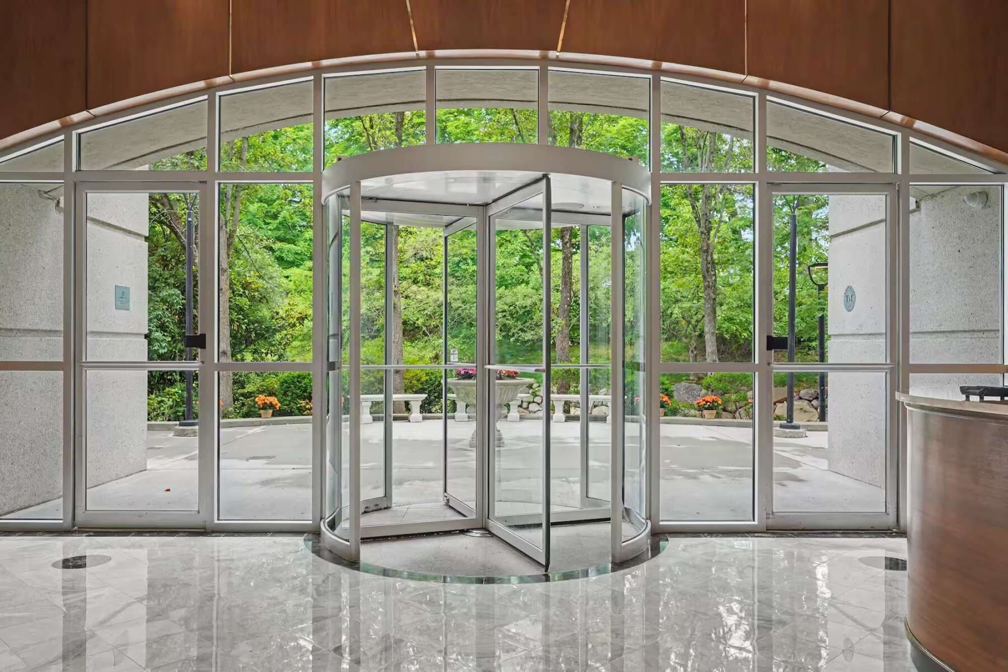A revolving door with glass panels, leading to an outdoor area with trees and a stone bench.
