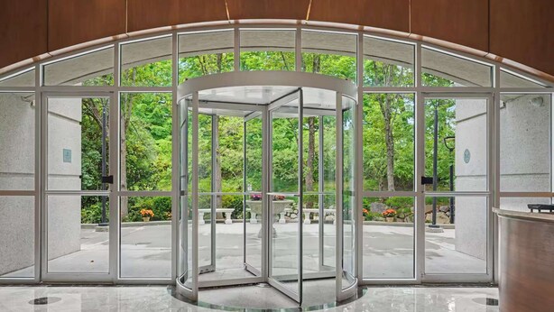 Une porte tournante à panneaux de verre, donnant sur un espace extérieur agrémenté d’arbres et d’un banc en pierre.