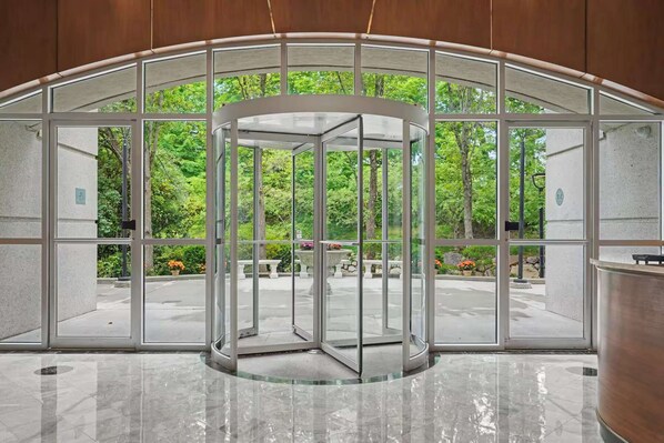 A revolving door with glass panels, leading to an outdoor area with trees and a stone bench.