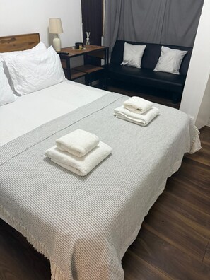 Una cama bien hecha con sábanas blancas y dos toallas dobladas, una mesita de noche de madera con una lámpara, un sofá oscuro con cojines blancos, y una cortina gris al fondo.