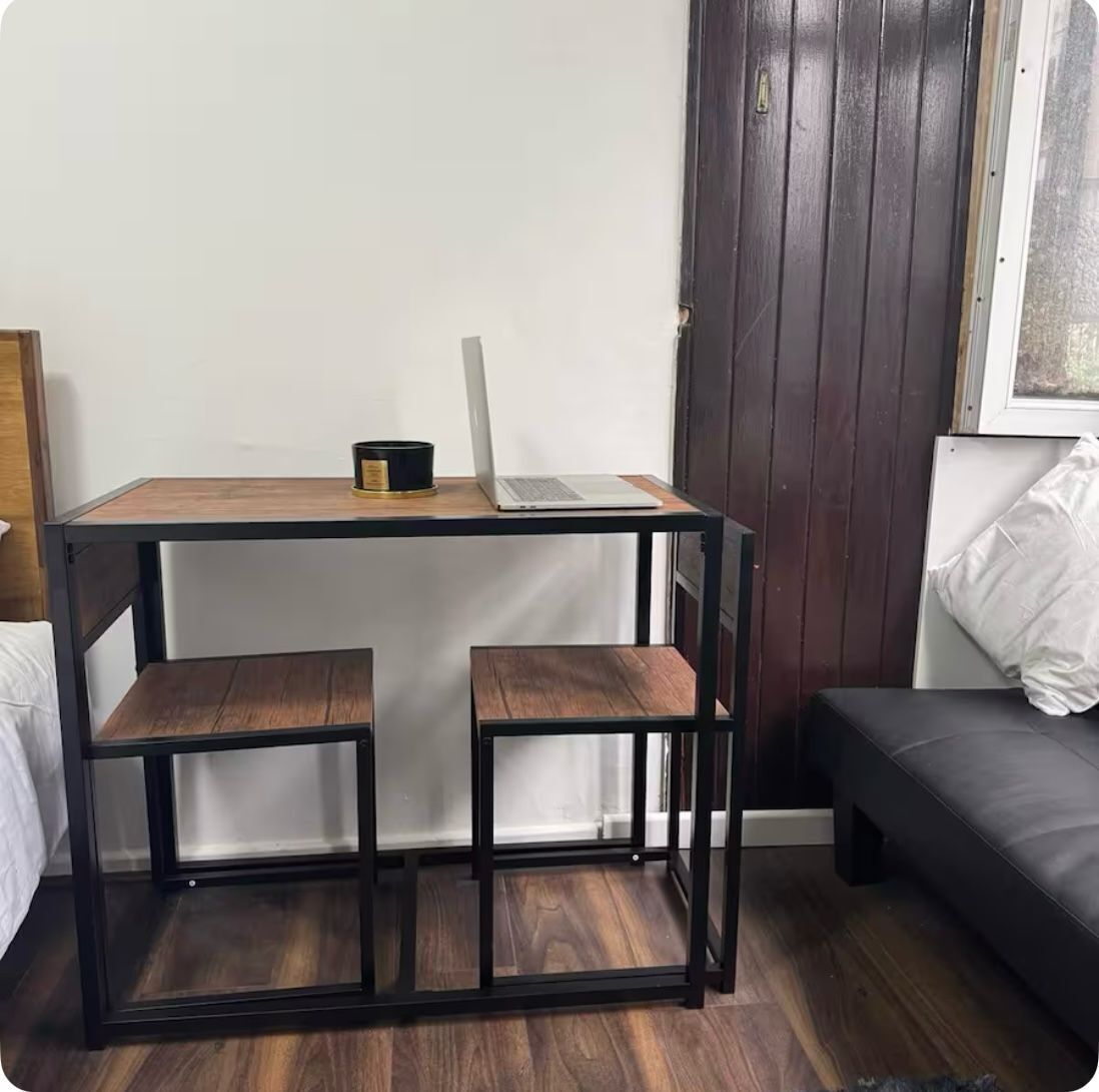 Une petite pièce avec un bureau en bois, deux chaises, un ordinateur portable et un canapé noir.