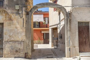 Une rue étroite, surmontée d’une arche en pierre, mène à un immeuble doté d’un balcon.