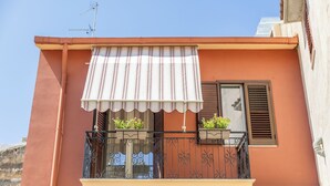 Ein Balkon mit gestreiftem Sonnensegel, ein Fenster mit Rollläden und Kübelpflanzen.