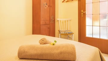 A neatly made bed with a rolled towel and a small flower, a wooden wardrobe, a white chair, and a door with a window.