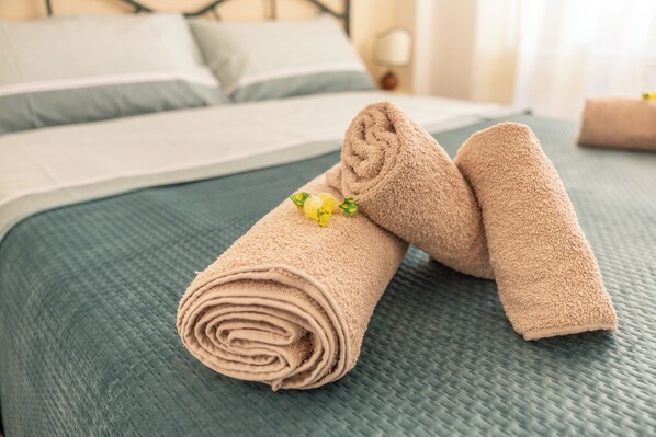 Una cama bien hecha con una colcha verde con textura y dos toallas enrolladas decoradas con una flor amarilla.