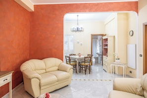 A living room with a beige sofa, a dining area with a table and chairs, and a kitchen in the background. - Casa del Sole - Menfi (Menfi)