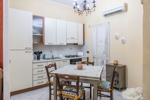 Une cuisine compacte avec des meubles blancs, une table à manger avec des chaises, un lustre et un climatiseur.