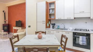 Uma cozinha compacta com armários brancos, uma mesa de jantar com cadeiras e uma pequena televisão sobre uma mesinha.