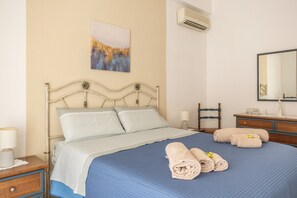 A neatly made bed with a blue bedspread and two rolled towels, a bedside table with a lamp, a chair, and a painting on the wall.