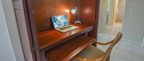 A wooden desk with a laptop and a desk lamp, a chair, and a tiled floor.