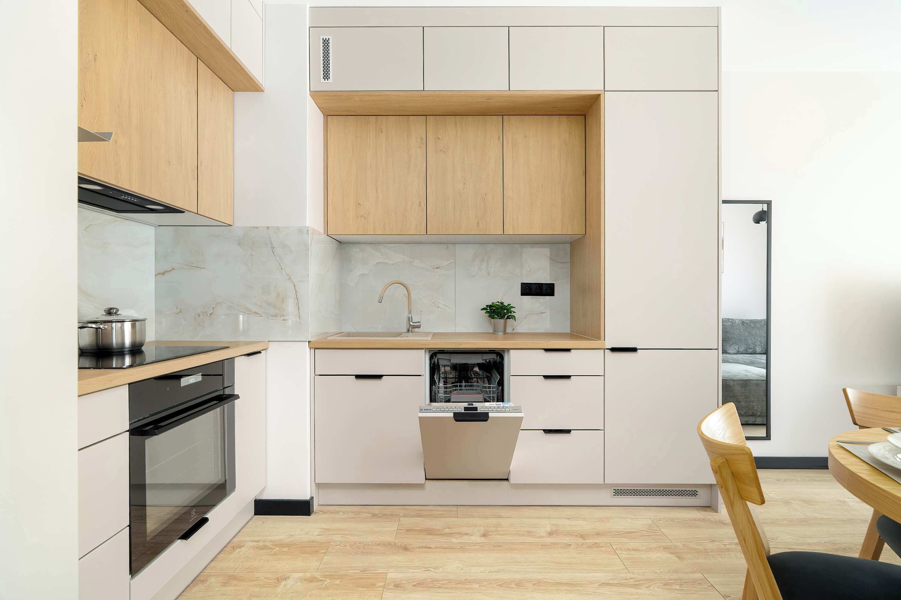 An open perspective of a well-lit kitchen area with two-tone cabinetry and marble-pattern backsplash. A small wooden dining set stands off to the side, maintaining an airy atmosphere.