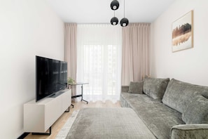 Ein modernes Wohnzimmer mit einem Flachbildfernseher, einem grauen Sofa, einem kleinen Tisch mit einer Pflanze und einem großen Fenster mit transparenten Vorhängen.