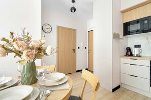 Una cocina moderna con microondas, cafetera y un armario de madera. Hay un comedor con una mesa lista para dos personas, un florero con flores y un reloj en la pared.