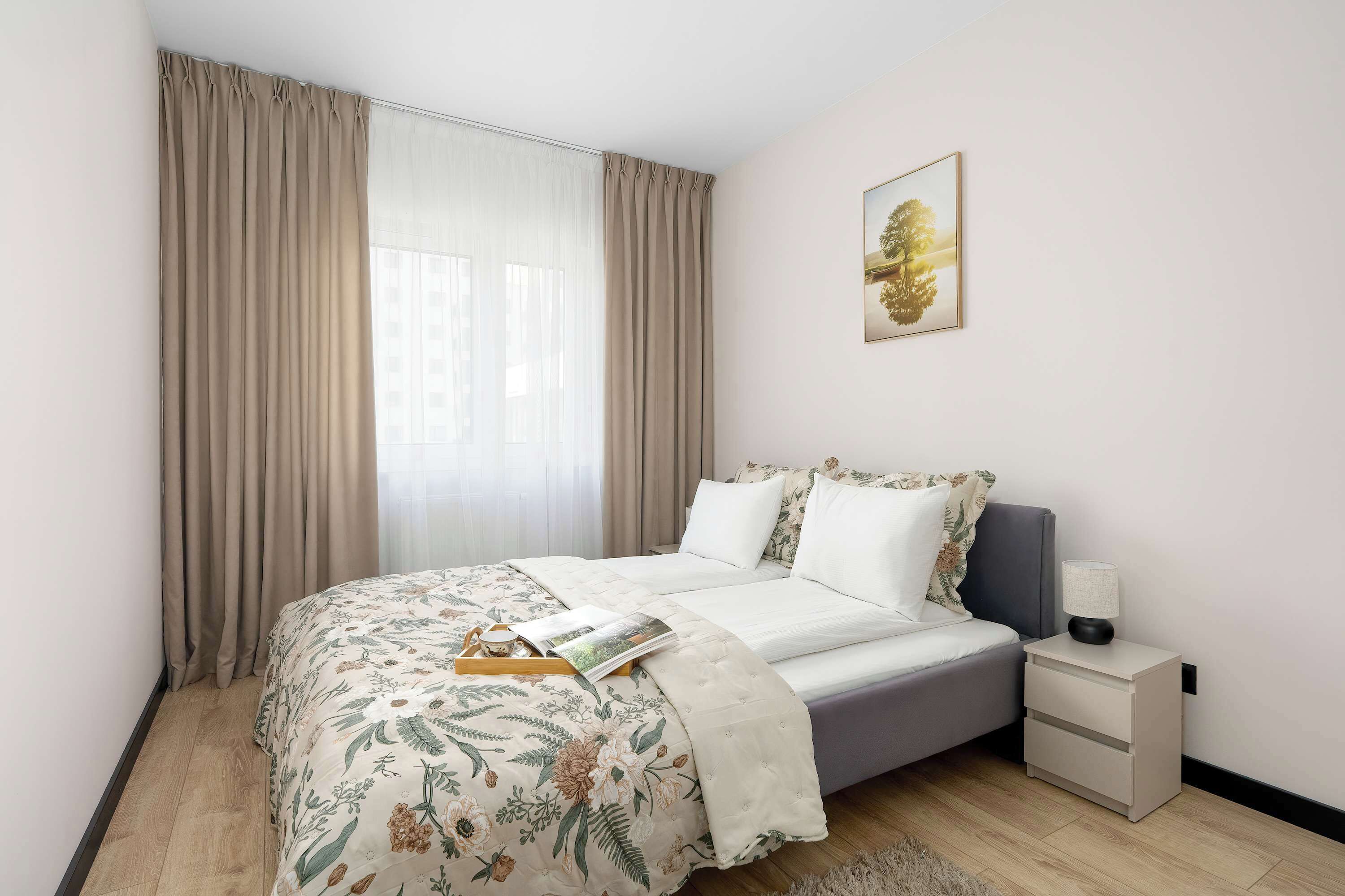 Un dormitorio con cama, mesita de noche, lámpara, un cuadro enmarcado en la pared y una ventana con cortinas.