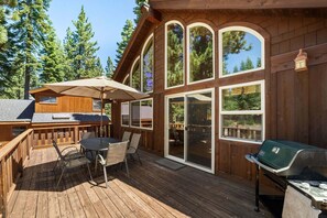 Eine Holzterrasse mit Tisch, Stühlen, einem Grill und großen Fenstern.