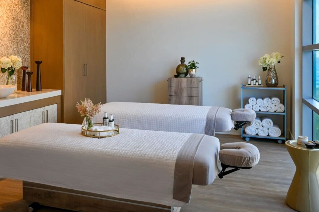 Une salle de spa équipée de deux lits, d’une petite table avec des serviettes et des articles de toilette, et d’une étagère contenant des serviettes.