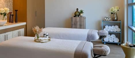 Une salle de spa équipée de deux lits, d’une petite table avec des serviettes et des articles de toilette, et d’une étagère contenant des serviettes.