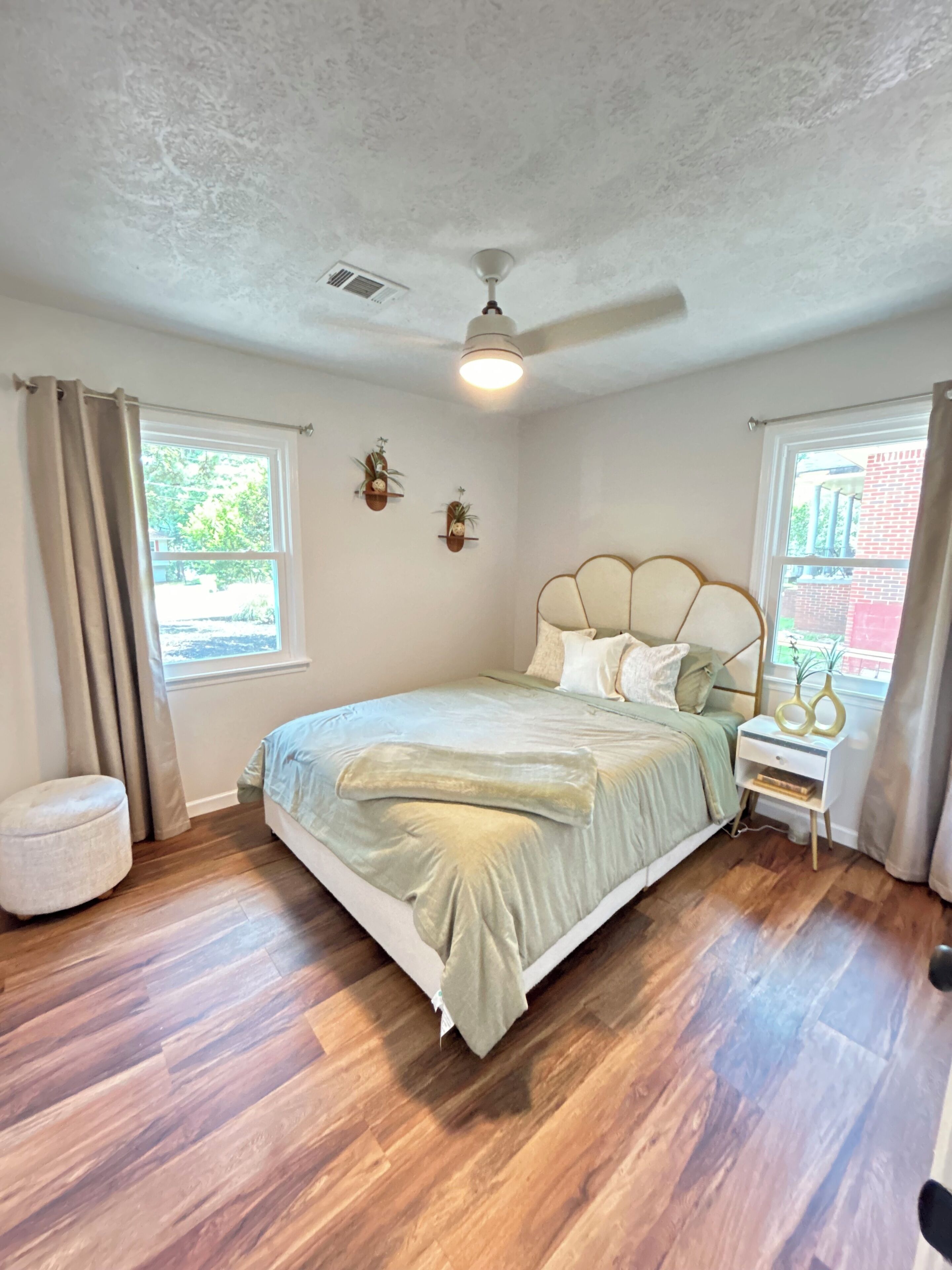Un dormitorio con cama, mesita de noche, ventilador de techo y dos ventanas con cortinas.