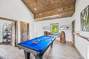 A pool table in a room with wooden beams on the ceiling, framed pictures on the wall, and a window with a view of greenery. - Stunning Mountain Views! 3BR Cabin w/ Hot Tub, Fire Pit & Games (Gatlinburg)