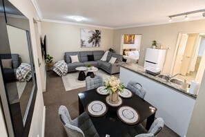 A modern living room with a grey sectional sofa, a dining table with placemats, and a kitchen area with a sink and countertop. - Kivuli | Walk to Elizabeth Quay • Pool • Parking (Perth)