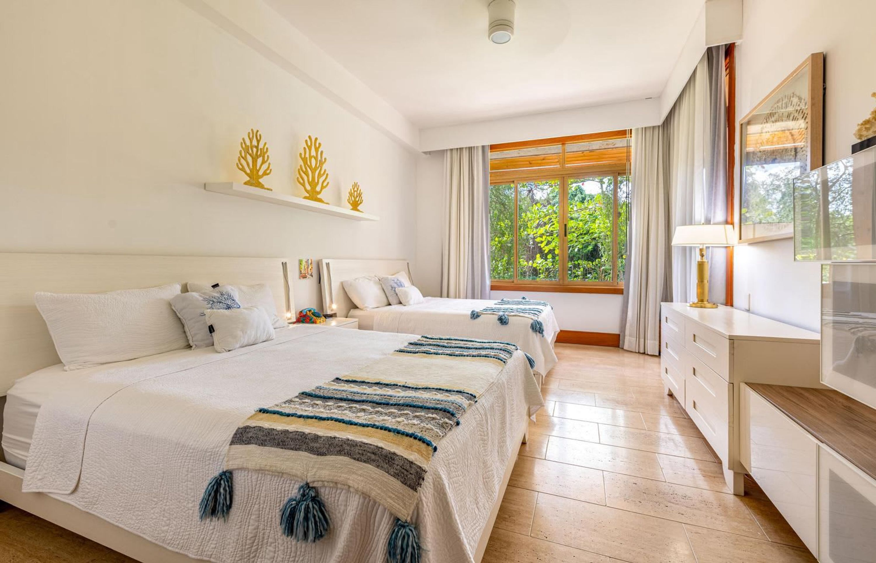 A hotel room with two beds, a large window with curtains, a wooden dresser, and decorative wall art.