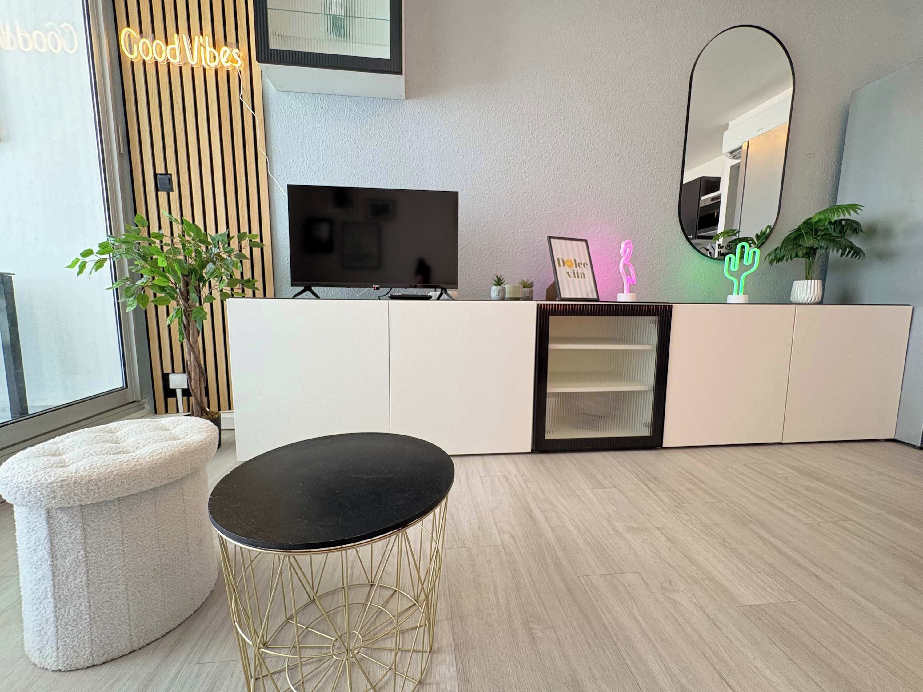 Un salon moderne avec un téléviseur, une table ronde à cadre doré, un pouf blanc et un grand miroir.