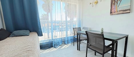 A room with a bed, a table with chairs, a view through a window, and a painting on the wall.