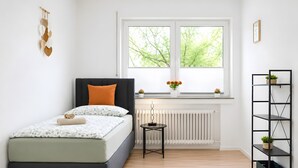 Un dormitorio con cama, un radiador, una ventana con vistas, una estantería con plantas y una obra de arte en la pared.