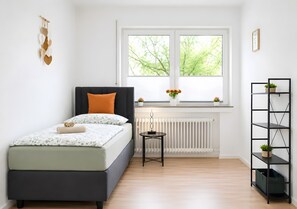 Ein Schlafzimmer mit einem Bett, einem Heizkörper, einem Fenster mit Blick, einem Regal mit Pflanzen und einem Wandbild.