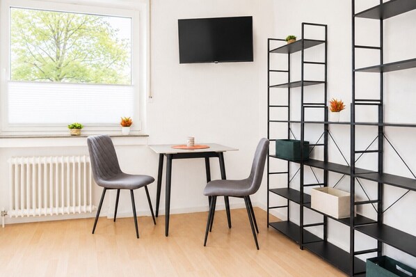 Une pièce avec un radiateur blanc, une fenêtre avec des stores, un téléviseur noir fixé au mur, une table blanche avec deux chaises grises et une étagère noire ornée de plantes.