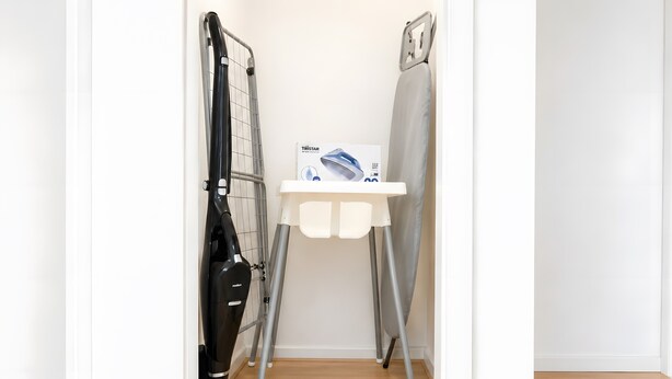 Habitación compacta con un aspirador, una plancha pequeña y una silla blanca.