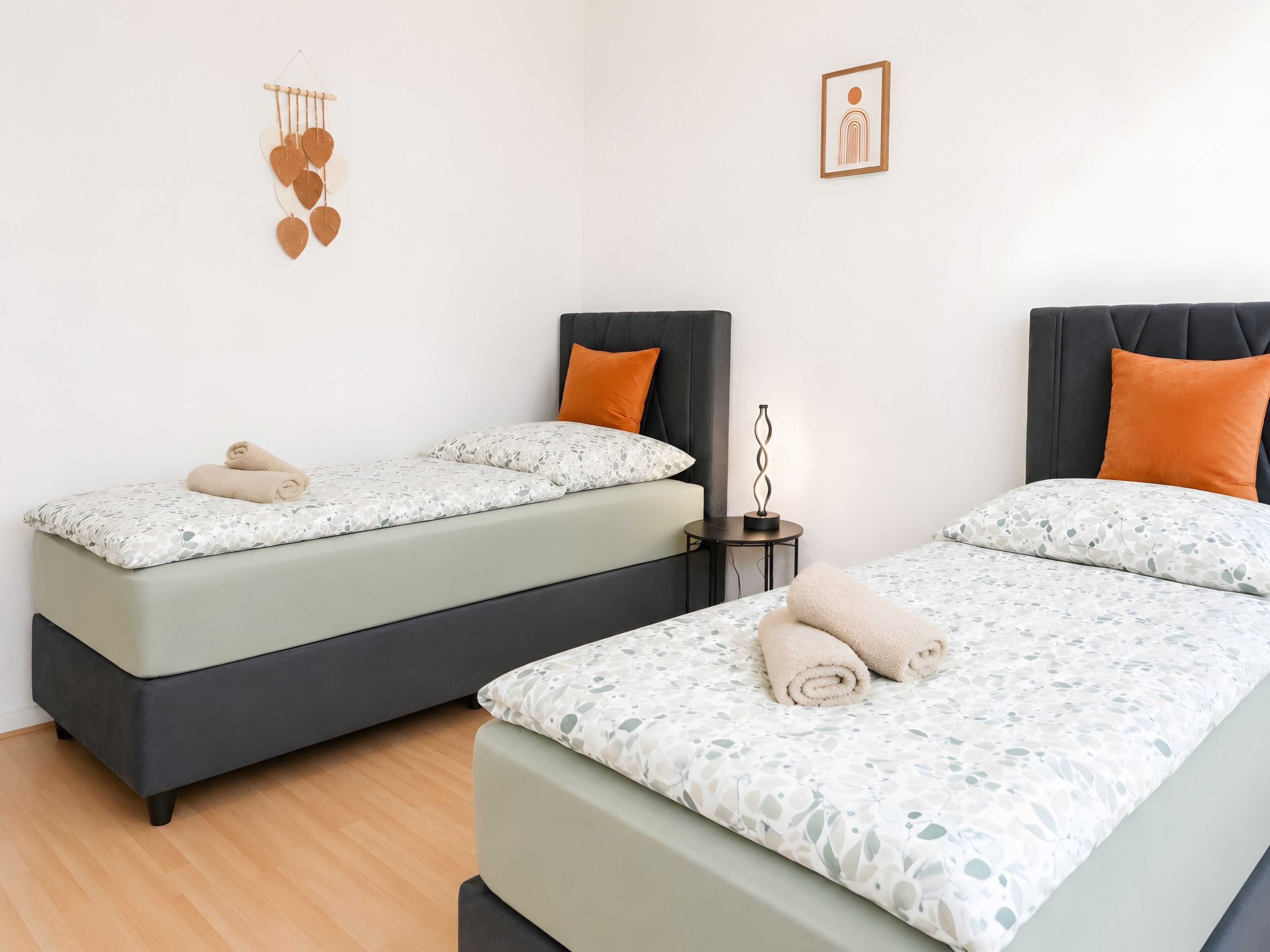 Ein Doppelzimmer mit zwei Betten, jeweils mit Kissen und einem gerollten Handtuch, einem Nachttisch mit Lampe sowie einer Wanddekoration.