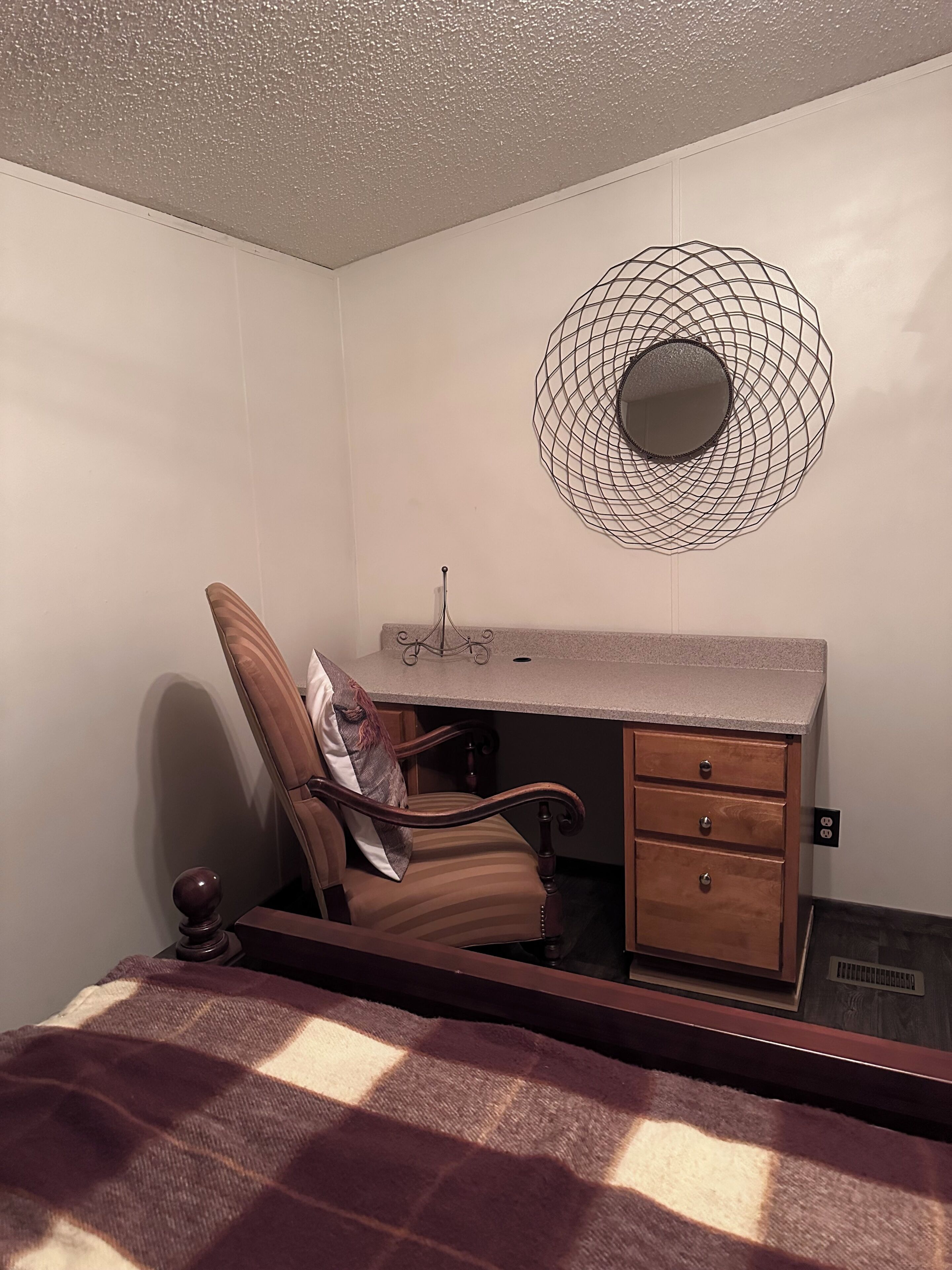 Un dormitorio con cama, una silla, un escritorio con cajones, un espejo en la pared y una lámpara en el techo.