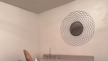 Un dormitorio con cama, una silla, un escritorio con cajones, un espejo en la pared y una lámpara en el techo.