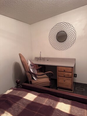 Un dormitorio con cama, una silla, un escritorio con cajones, un espejo en la pared y una lámpara en el techo.
