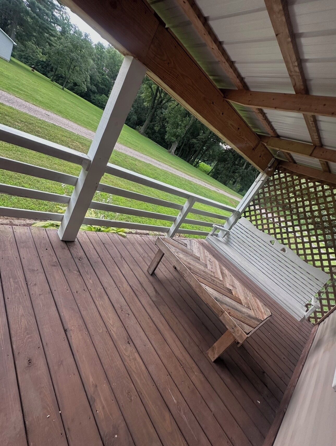 Una terraza de madera con barandilla blanca, un espacio cubierto con techo metálico y vistas a un campo césped y árboles al fondo.