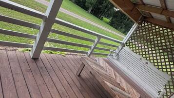 Una terraza de madera con barandilla blanca, un espacio cubierto con techo metálico y vistas a un campo césped y árboles al fondo.