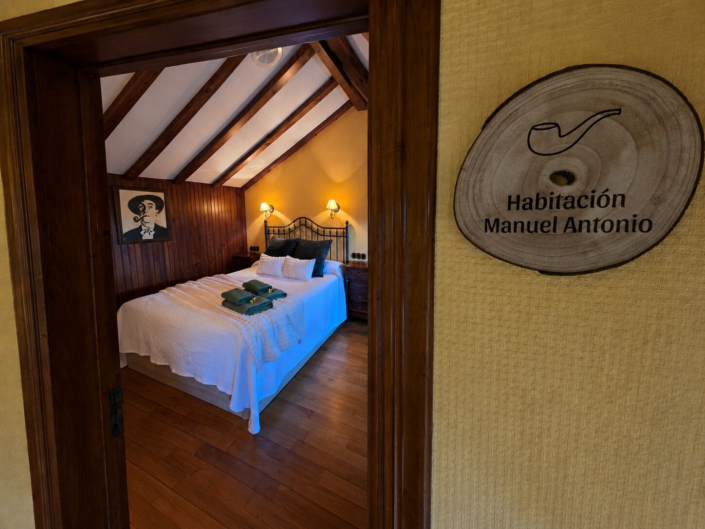 A bedroom with a wooden ceiling, a bed with white bedding, and a wall-mounted sign reading 'Habitación Manuel Antonio'.