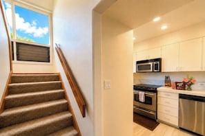 A modern kitchen with stainless steel appliances, a microwave, and a stove. There is a window above the staircase, and a small table with a vase and a box of Hawaiian Host cookies. - Kapalua Ridge | 2 Ocean View Villas, Sleeps 12 | Car Incl. w/ 6+ Nights | ML-4253 by KBM (Lahaina)