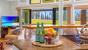 A modern hotel room with a TV displaying a tropical beach scene, a bar area with bottles and fruit, and a view of a golf course. - Kapalua Ridge | 2 Ocean View Villas, Sleeps 12 | Car Incl. w/ 6+ Nights | ML-4253 by KBM (Lahaina)