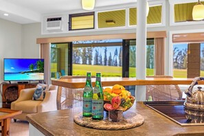 A modern hotel room with a TV displaying a tropical beach scene, a bar area with bottles and fruit, and a view of a golf course. - Kapalua Ridge | 2 Ocean View Villas, Sleeps 12 | Car Incl. w/ 6+ Nights | ML-4253 by KBM (Lahaina)