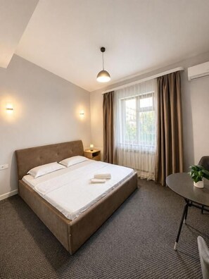 Une chambre d’hôtel avec un lit simple, une table de chevet, une petite table avec une plante et une fenêtre avec des rideaux.