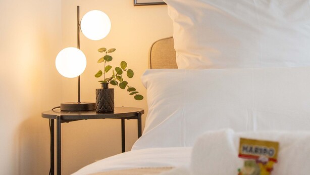 En snyggt bäddad säng med vita sängkläder, ett litet nattduksbord med en lampa och ett inramat konstverk på väggen.