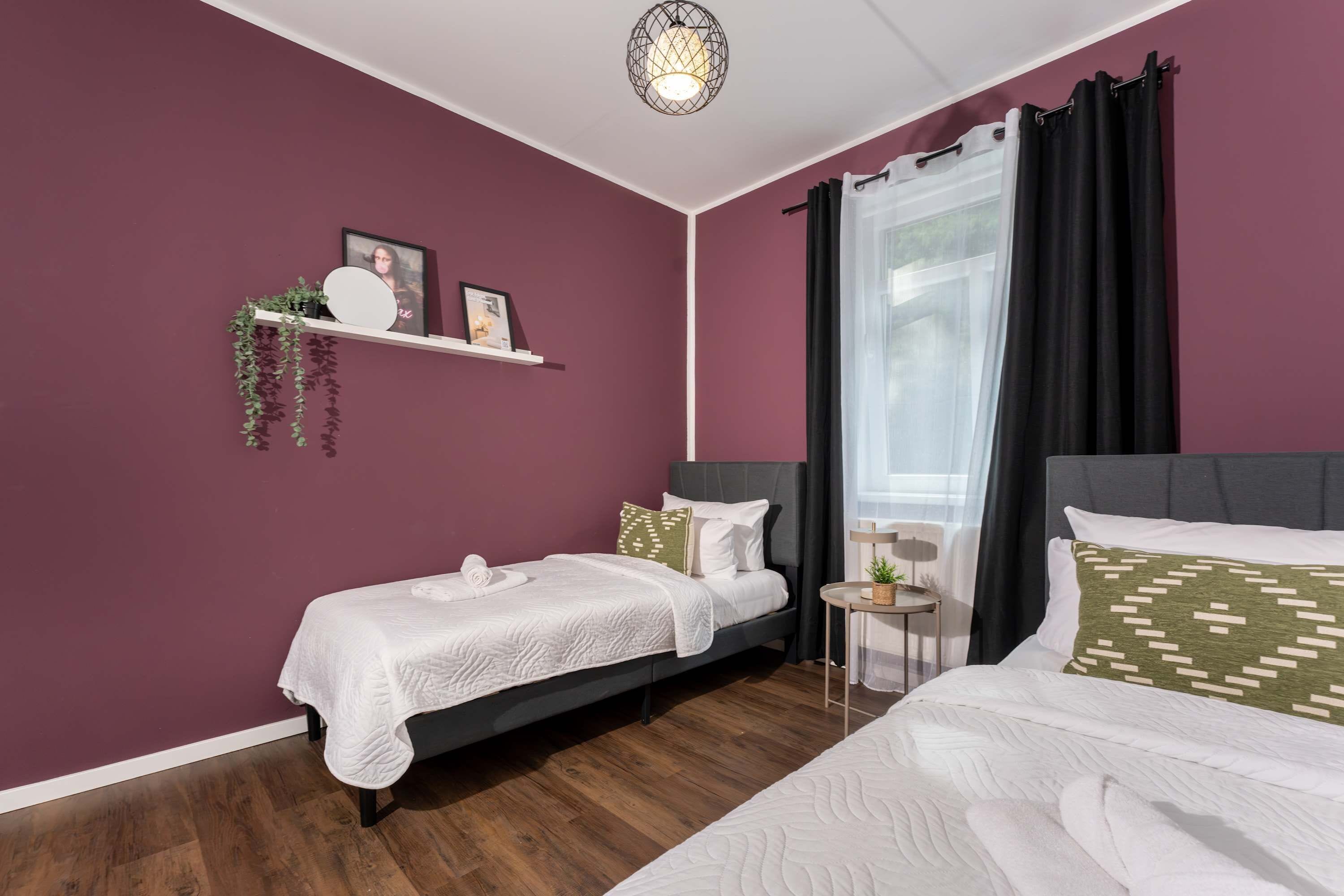 Un dormitorio con dos camas, una ventana con cortinas, una mesita de noche y un estante en la pared con plantas y fotos.