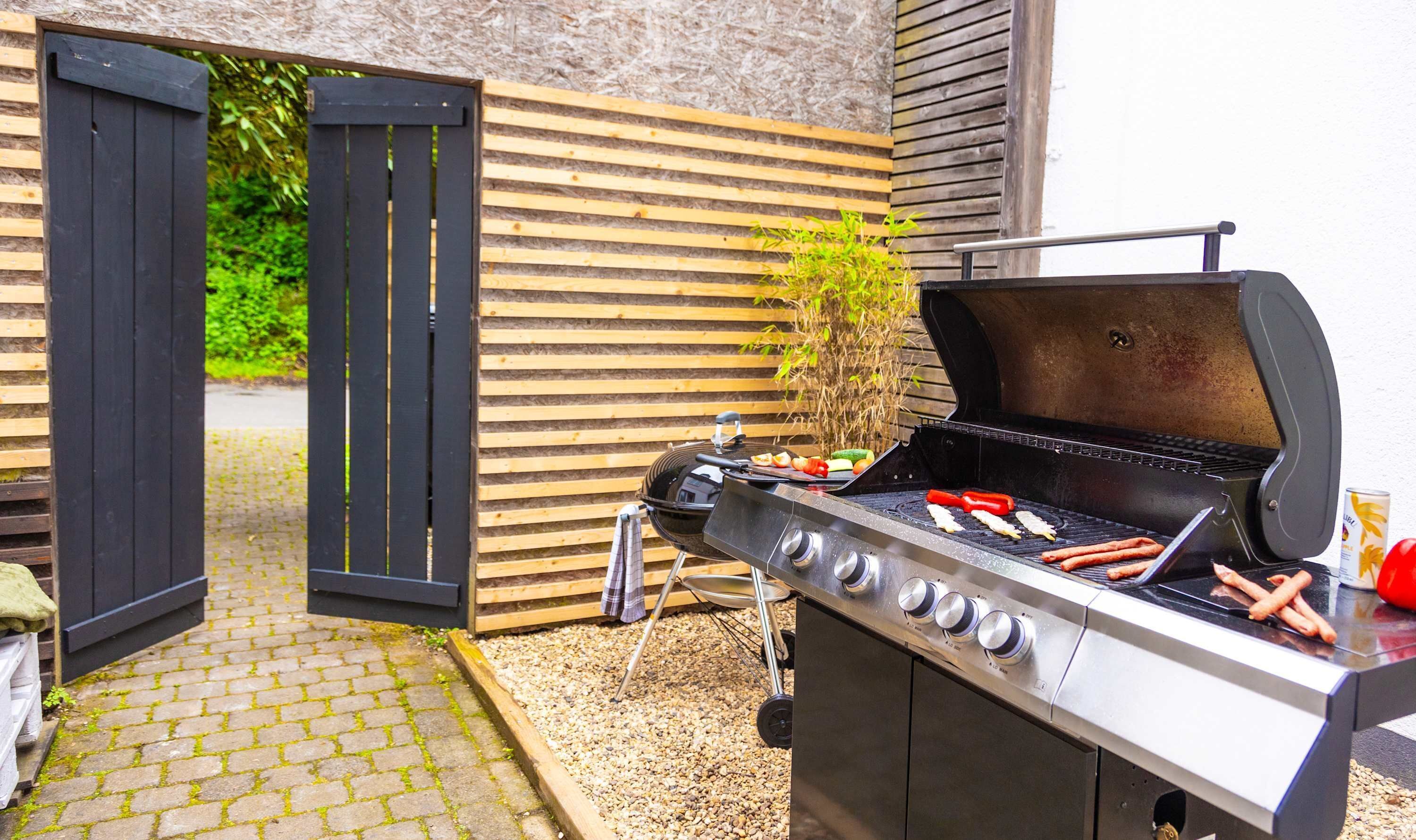 An outdoor grill with sausages and vegetables, a wooden fence, and a paved pathway.