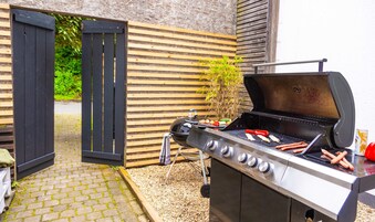 Una parrilla al aire libre con salchichas y verduras, una valla de madera y un camino pavimentado.