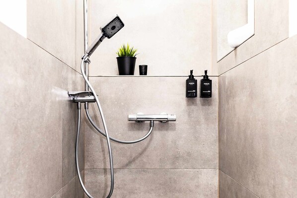 A modern bathroom with a handheld showerhead, a small potted plant, and two bottles of shampoo and conditioner on a shelf.