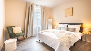 Un dormitorio con cama, una silla, una mesita de noche y una ventana con cortinas.